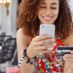 Woman with curly hair smiles as she looks at her phone in her left hand and holds a credit card in her right hand.