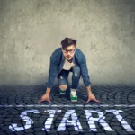 Man in jeans and sneakers with a denim top squats on cobblestones in front of a line that says START