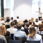 A group of people sits in an conference space facing a speaker at the front of the room.