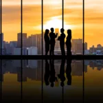 Four people are standing in an empty building in front of a large window. In the background there is a city skyline with the sun setting behind it.