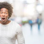 A man wearing a white sweater is holding a blue alarm clock. He has a shocked expression on his face. The background behind him is blurred out.