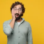 Bearded man wearing a long-sleeved green, button-down shirt holds his hand to his cheek in surprise