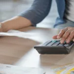 Hands lay on wooden desk with one hand holding a piece of paper and another punching in numbers on a calculator.