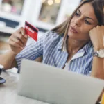 A woman is sitting in a coffee shop at a table looking at her credit card. On her table there is a cup of coffee, a laptop, and a green drink in a mason jar.