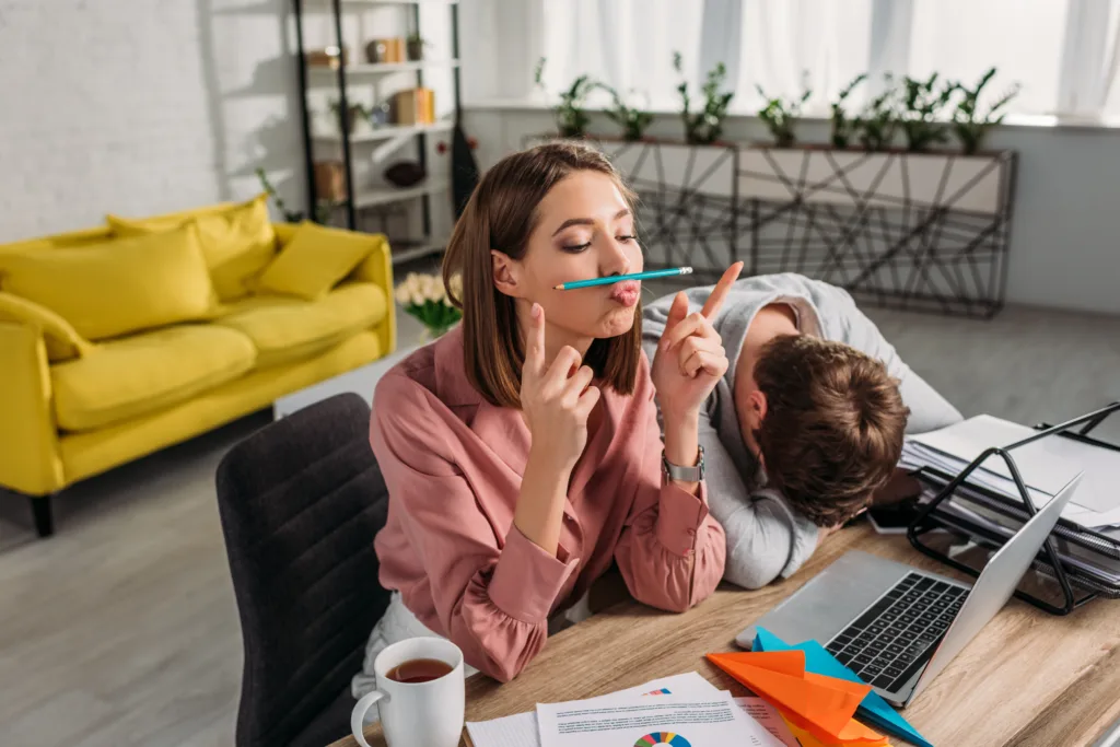 Woman procrastinates by trying to balance a pencil between her nose and lips while a man sits with his head in his arm next to her.