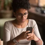 A woman wearing glasses, a silver necklace, a pink shirt, a red bracelet, and a wrist watch is sitting down staring at a smartphone. The background behind her is blurry.