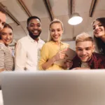 A group of men and women are surrounding a laptop. They are all looking at the screen smiling. They are at a desk in an office space.