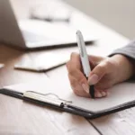 Someone is writing with a black and white pen on a piece of paper attached to a clipboard. The person has a gray suit on and there is a silver laptop and some scattered documents in the background.