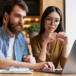 A man and woman looking at an open laptop. Behind them, is a coffee shop with ambient lighting.