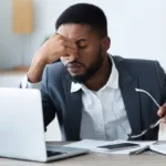 A man in a suit jacket and white dress shirt holds his eyeglasses in front of his laptop and holds his head in his hands while looking disappointed.