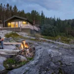 Photograph of person sitting on mountain reading and relaxing in front of a fire with a brightly lit cabin in the background