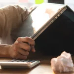 A person with their face down on a desk holding their laptop over their head. In front of them there is a calculator and a crumpled up piece of paper.