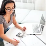 Woman with jeans and grey top with black rim glasses looks at a calculator and papers with a red pen while her laptop is set to the side.