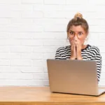 Blonde woman with hair tied in bun in black-and-white striped top and mustard-rimmed glasses covers her mouth with her hands in shock while a laptop sits open in front of her.