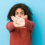 Woman with curly brown hair and orange sweater holds her hands in an X in front of her