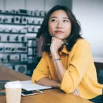 Woman in yellow blouse sits in a coffee shop with her chin in her hand and looks off in the distance contemplatively