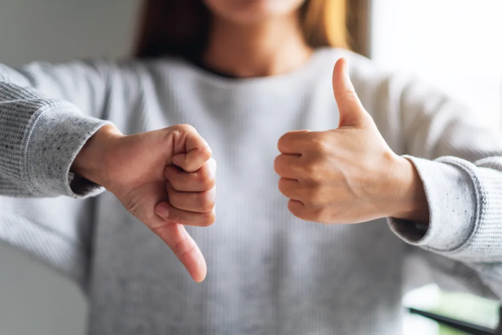 Person in grey crewneck sweatshirt is out of focus with their hands in focus in the foreground with one in a thumbs up and one in a thumbs down.