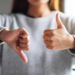 Person in grey crewneck sweatshirt is out of focus with their hands in focus in the foreground with one in a thumbs up and one in a thumbs down.