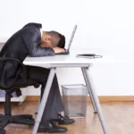 Man in suit sits in a rolling chair at a metal desk with his head laying face down on his laptop.