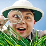 Man in safari hat with magnifying glass up to his eye is on the ground and looking in the grass.