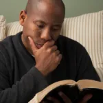 Man leaning against yellow-and-white striped pillow, reading a book, and holding his hand to his chin in contemplation