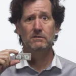 Man has a look of concern and sadness on his face as he raises up a tiny one-dollar bill pinched between his fingers.