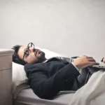 Man wearing glasses and a suit lies in bed with his laptop on his chest looking exhausted