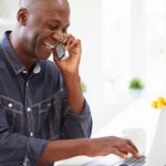 Bald Black man in a denim top laughs while talking on the phone and typing on his laptop.