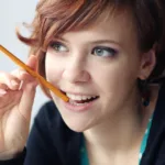 Redheaded woman looks to her right with a half smile and the eraser part of the pencil perched on her lips in contemplative thought.