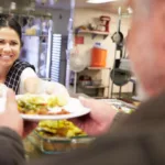 Woman at a food kitchen wearing serving gloves hands over a plate of food to a a man on the other side of the counter.