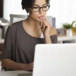 Woman in brown shirt and glasses sits in front of open laptop