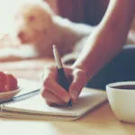 Woman's hand is shown writing on a notebook with a pen next to a cup of black coffee with a dog in the background