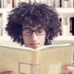 Man with curly dark hair and wearing glasses reads a book in front of a bookcase