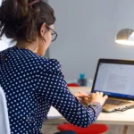 Looking over the shoulder of a woman in a navy polka dress, with glasses on, works on her laptop computer.