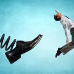 A giant black business shoe is at the end of a spring and booting a man in a business suit against a blue background.