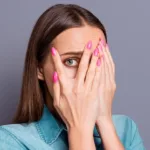 Woman in denim top and Barbie-pink nails covers her face with her hands with one eyeball peeking through one hand.