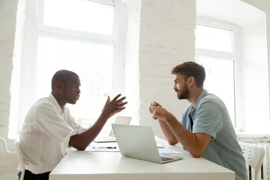 Two men face each other across a table with a laptop in front of them as they discuss work