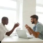 Two men face each other across a table with a laptop in front of them as they discuss work