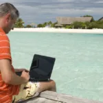 Man in orange and white striped shirt and shorts sits on deck overlooking a beach typing on a laptop