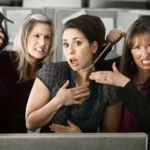 Three women are all dressed in black outfits as one on the left threateningly holds a heel up to the one in the middle while the woman on the right pulls at the middle woman's pearl necklace.