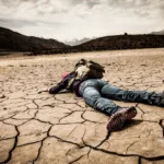 Person is sprawled across a cracked dirt ground with mountains in the distance.