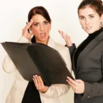 Two women, one in a white blazer and one in a black blazer are looking at a portfolio with shocked expressions, one holding a hand to her head and one holding a hand out as if to say, "what is this?"