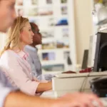 Three workers sit side-by-side working on the computer; the man in the foreground is blurred and focus is on blonde woman in pink shirt.