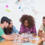 A group of young professionals in casual dress sit around a table with water glasses looking at notes on paper while a whiteboard sits behind them covered in writing and sticky notes.