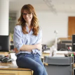 Red-headed woman in blue button-down shirt and jeans wearing glasses smiles as she sits with her arms crossed on top of a desk within a creative office space.