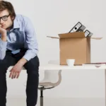 Man sits with his head resting on his hand which is resting on his knee while the desk next to him has a box packed with office belongings.