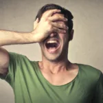 Man in green t-shirt holds his right hand over his face with his mouth open in a scream.