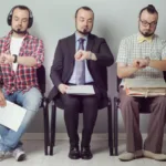 Three men sit side-by-side in chairs all looking at their watches. One is in a plaid shirt and jeans, one in a suit, and one in a button-down shirt with bow tie.