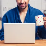 Bearded man in blue bathroom sits in front of his laptop holding a coffee mug