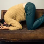 Woman in brown sweater and jeans leans forward while sitting on a sofa holding her head in her hands